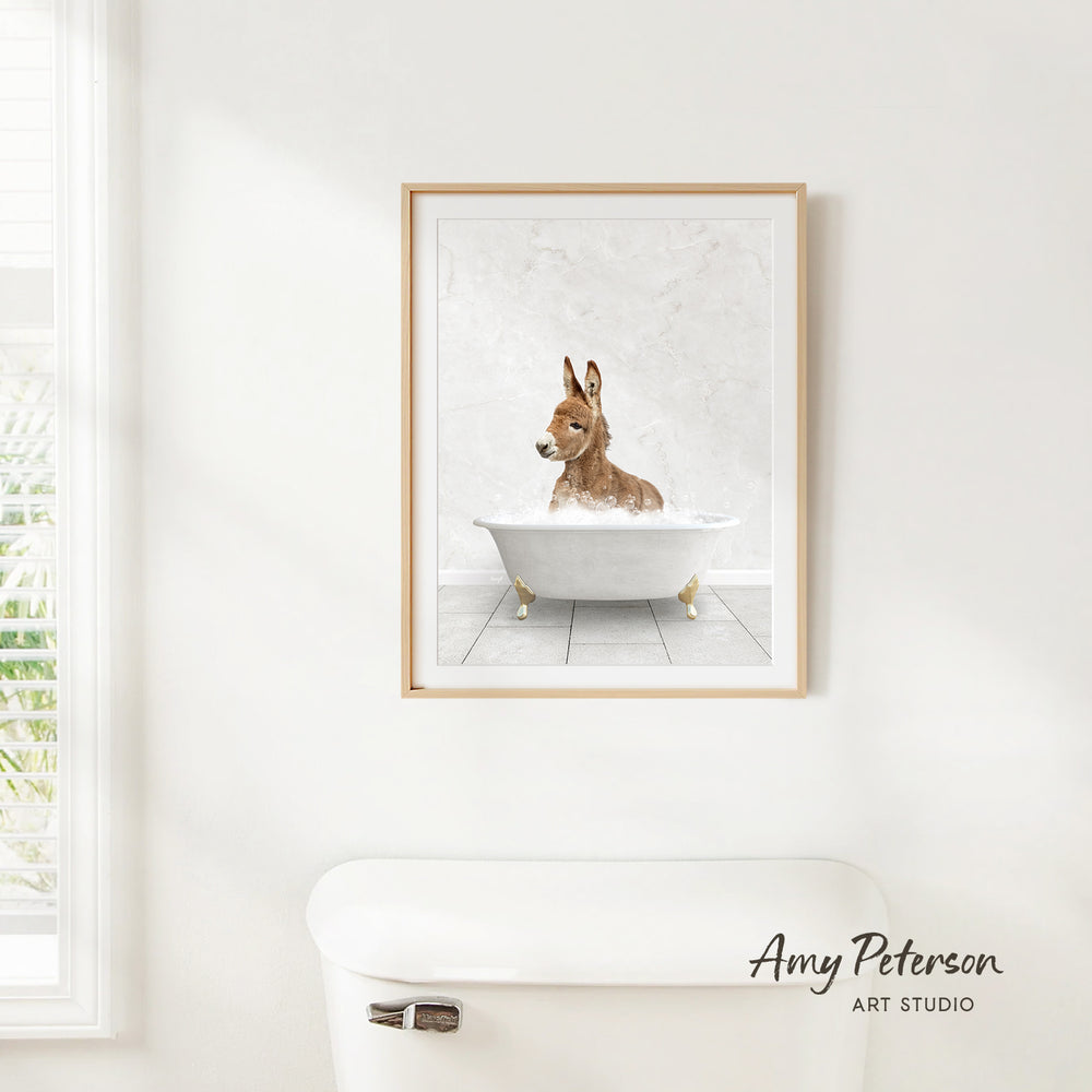 A framed print of a donkey sitting in a bathtub is hanging on a white wall.