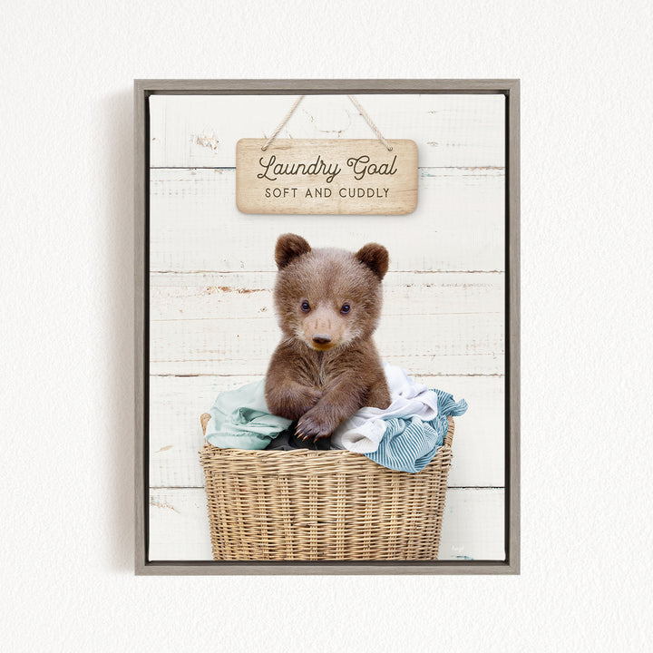 A cute brown bear sitting in a wicker laundry basket, with a wooden sign above that says "Laundry Goal Soft and Cuddly".