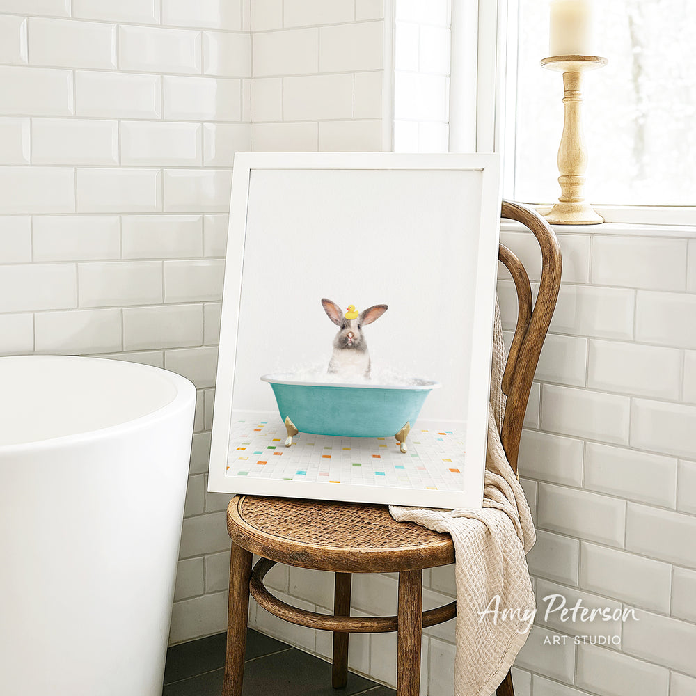 A framed image of a rabbit sitting in a blue bathtub, surrounded by colorful tiles, is displayed on a wooden chair in a bathroom setting.
