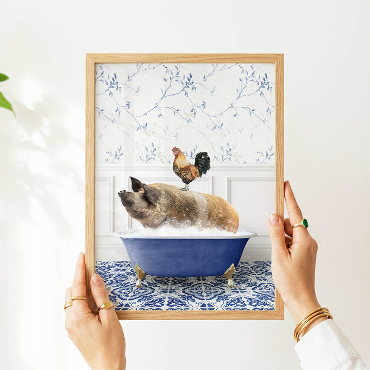 A person's hands are holding a framed image that depicts a pig sitting in a blue bathtub, with a rooster perched on its back.