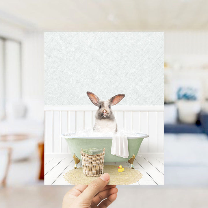 A hand holds up a framed image of a rabbit sitting in a bathtub filled with bubbles, surrounded by a basket of toys and a yellow rubber duck.