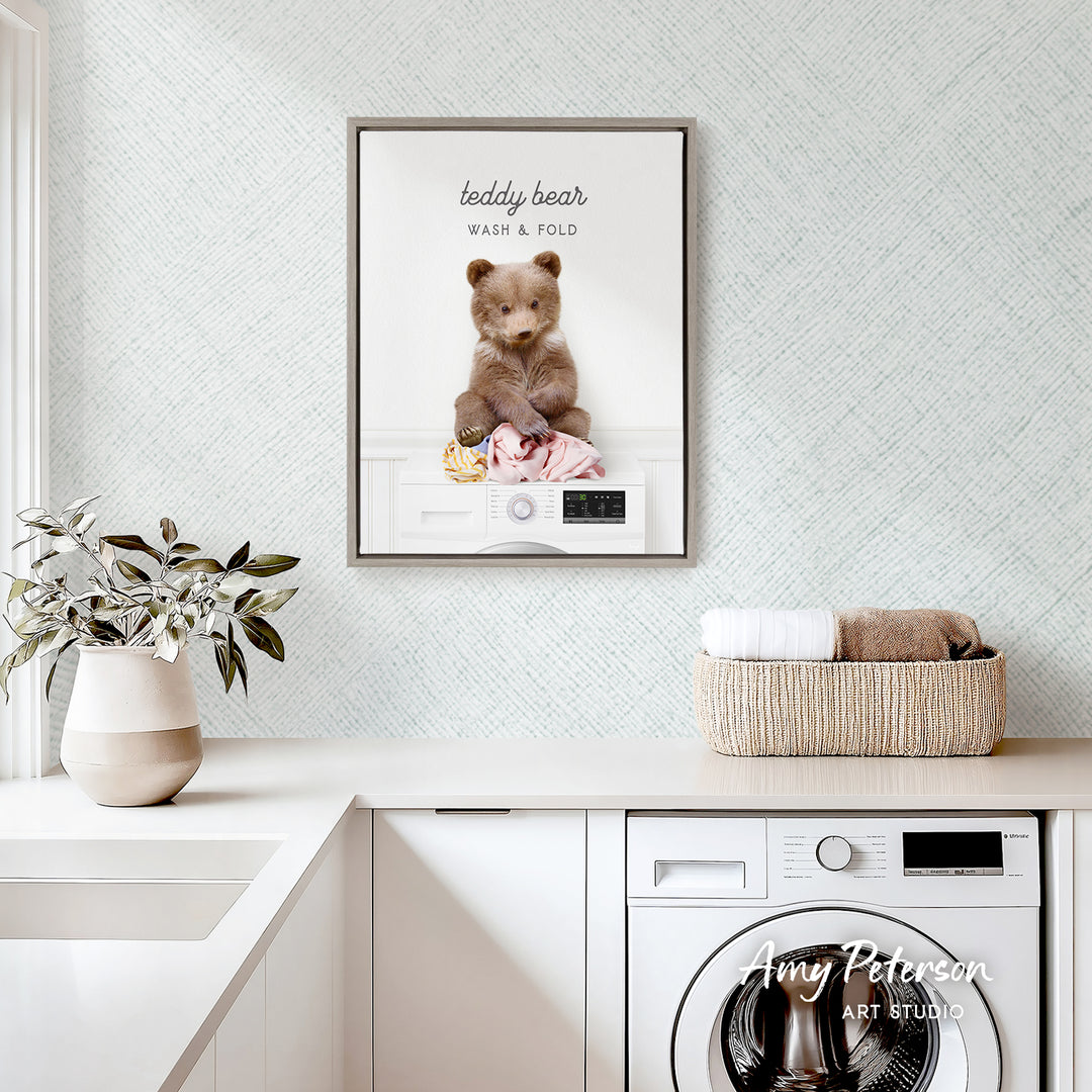 a teddy bear sitting on a washing machine, with a framed picture of the teddy bear above it. The teddy bear is wearing a pink outfit and appears to be in the process of being washed. The teddy bear is sitting on a white towel, and there is a basket of clothes nearby. The backdrop is a light blue wall with a patterned design.