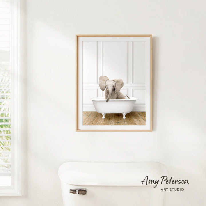 A framed image of a baby elephant sitting in a white bathtub, with a window and a toilet visible in the background.