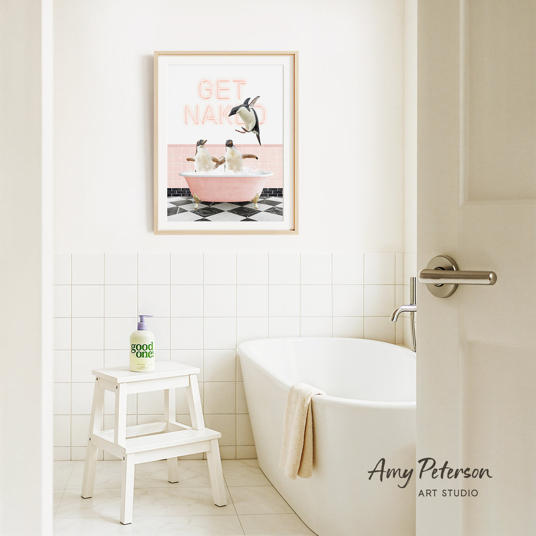 A bathroom with a white bathtub, a stool, and a framed poster of penguins in a pink bathtub. The poster has the text "GET NAKED" written on it.