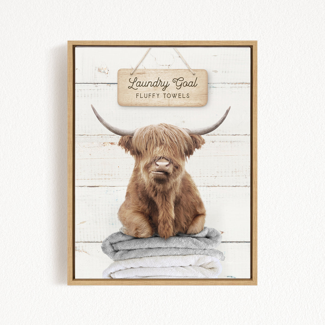 A framed image of a brown cow with long horns sitting on a stack of fluffy towels, with a sign above that says "Laundry Goal Fluffy Towels".