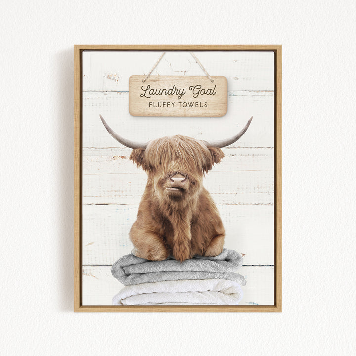 A framed image of a brown cow with long horns sitting on a stack of fluffy towels, with a sign above that says "Laundry Goal Fluffy Towels".
