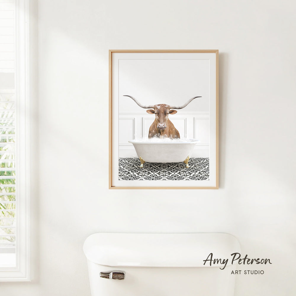 A framed image of a bull with large horns sitting in a bathtub, with a patterned tile floor beneath it.