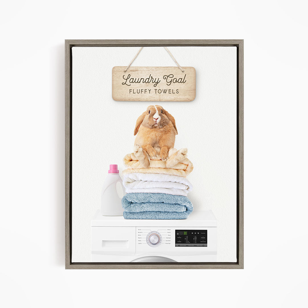 A wooden sign with the text "Laundry Goal Fluffy Towels" hangs above a white washing machine with a pile of folded towels on top.