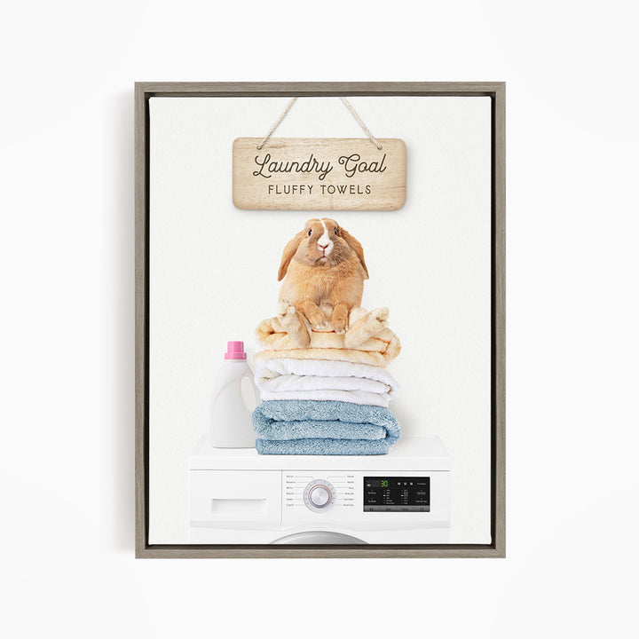 A wooden sign with the text "Laundry Goal Fluffy Towels" hangs above a white washing machine with a pile of folded towels on top.