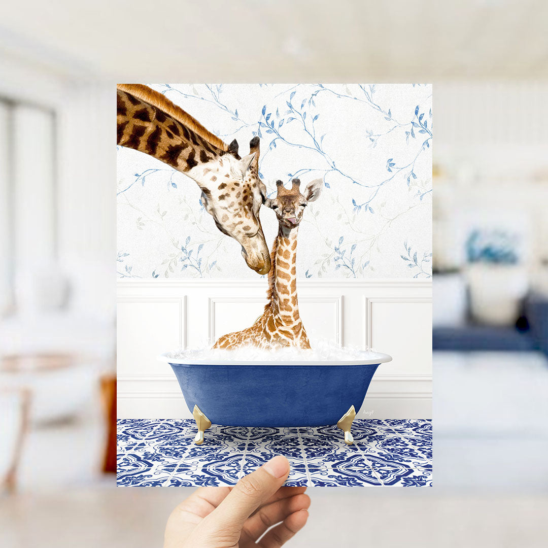 A hand holds up a framed image of two giraffes sitting in a blue bathtub, with one giraffe appearing to be bathing the other.
