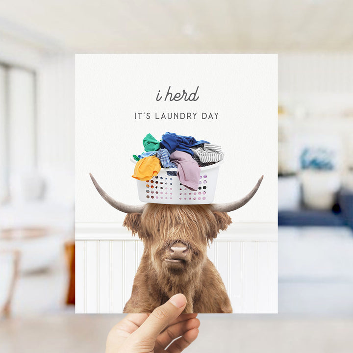 A person is holding up a card with a picture of a Highland cow wearing a laundry basket on its head, with the text "It's laundry day" written above it.