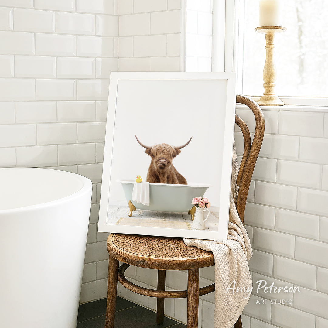 A framed image of a brown cow sitting in a white bathtub, with a towel and flowers nearby, is displayed on a wooden chair in a bathroom setting.