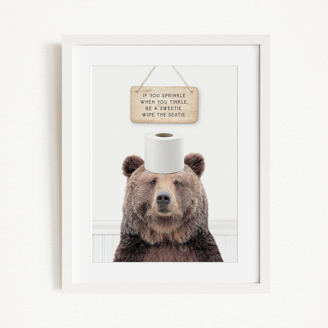 A framed image of a brown bear sitting on a roll of toilet paper, with a wooden sign above it that reads "If you sprinkle when you twinkle, wipe the seagull".