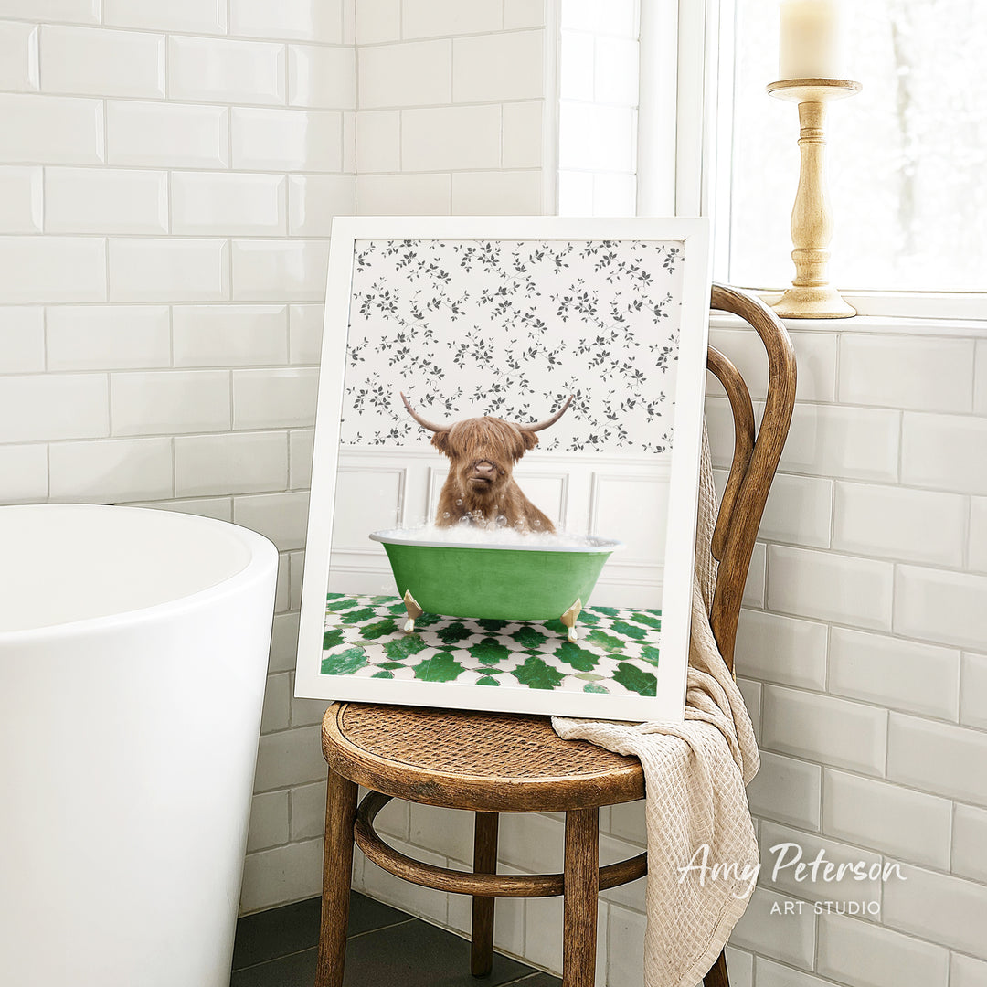 A framed image of a Highland cow sitting in a green bathtub, surrounded by a floral pattern, is displayed on a wooden chair in a bathroom setting.