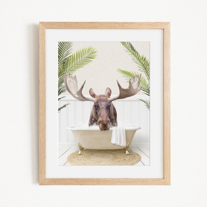 A framed image depicting a moose with large antlers sitting in a bathtub, surrounded by tropical plants.