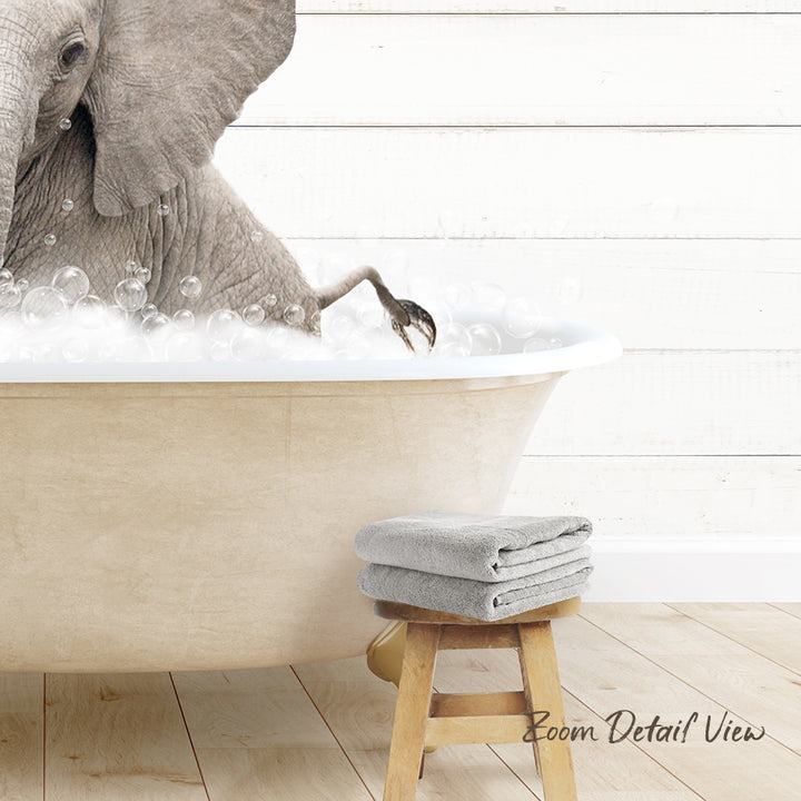 A large elephant is sitting in a bathtub filled with bubbles, with a towel and stool nearby.