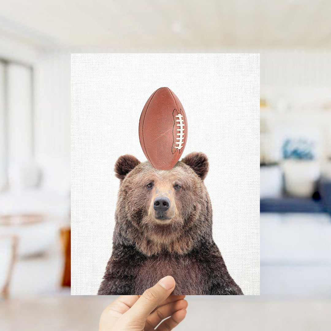 A hand holds up a square print featuring a bear with a football on its head, set against a white background.