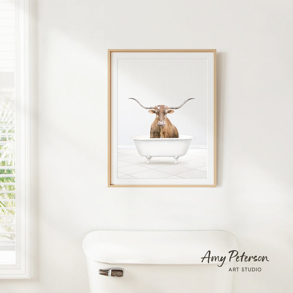 A framed image of a bull with horns sitting in a bathtub is hanging on a white wall.