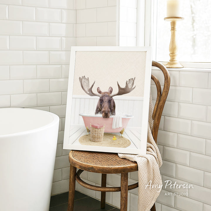 A framed image of a moose sitting in a pink bathtub, with a towel and a basket nearby, is displayed on a wooden chair in a bathroom setting.