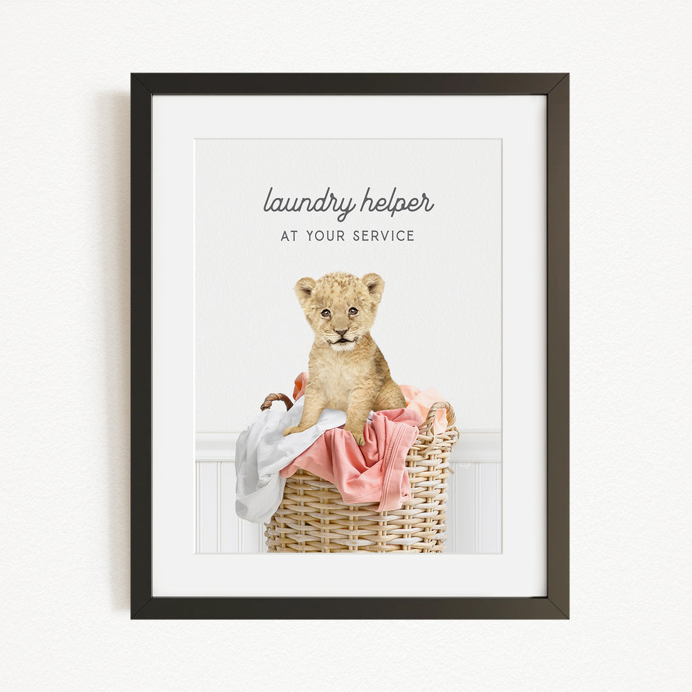 A framed image of a young lion cub sitting in a wicker basket filled with clothes, with the text "laundry helper at your service" written above the image.