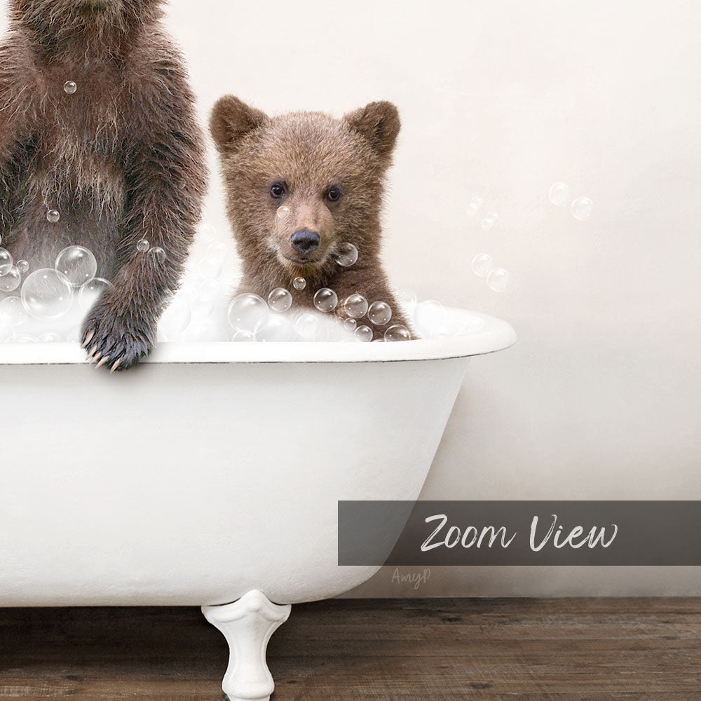 A baby bear sitting in a bathtub filled with bubbles, with a white bathtub and a wooden floor visible in the image.