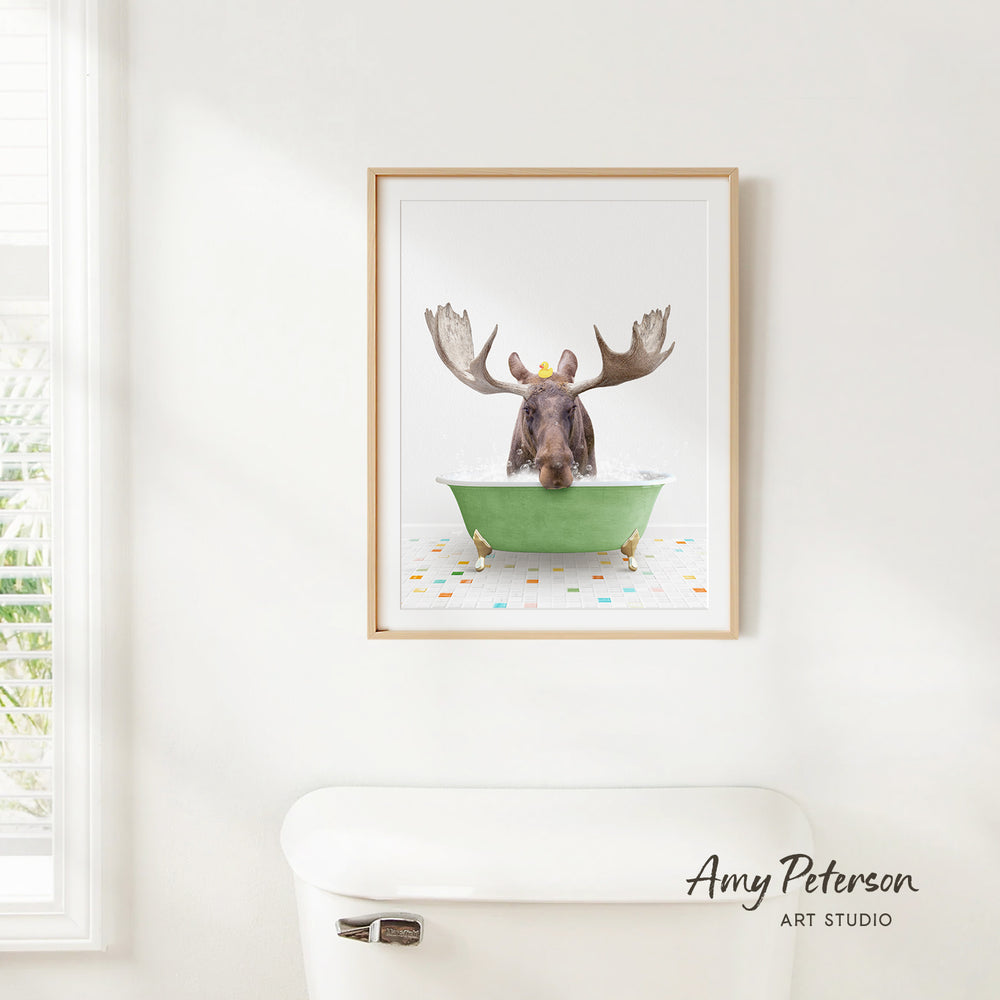 A framed image of a moose sitting in a green bathtub, with its large antlers raised, is hanging on a white wall.