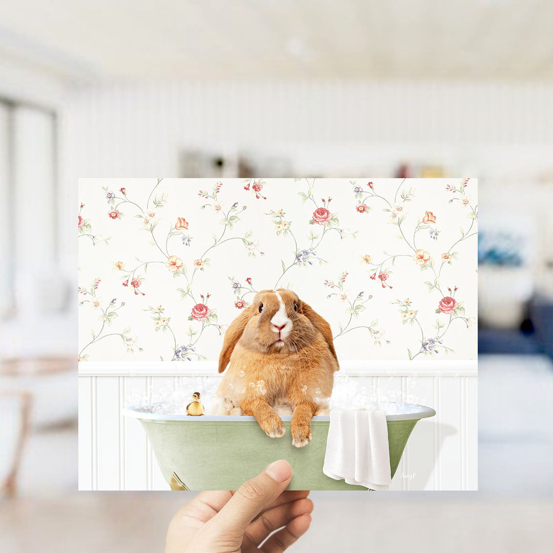A hand holding a framed image of a brown rabbit sitting in a green bathtub filled with water, surrounded by a floral patterned wallpaper.