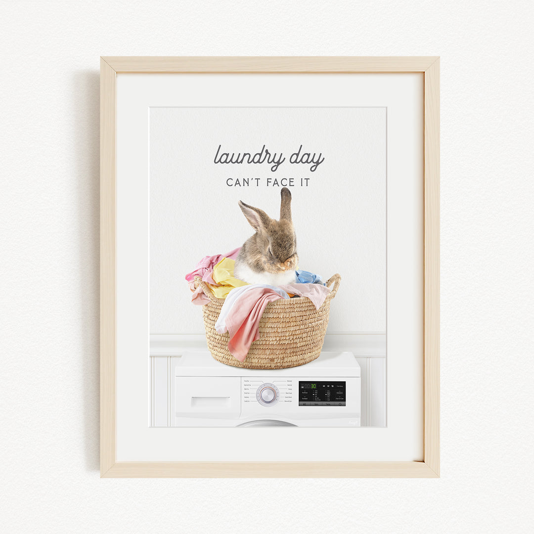 A framed image depicting a rabbit sitting in a woven laundry basket filled with clothes, with the text "laundry day can't face it" above the basket.