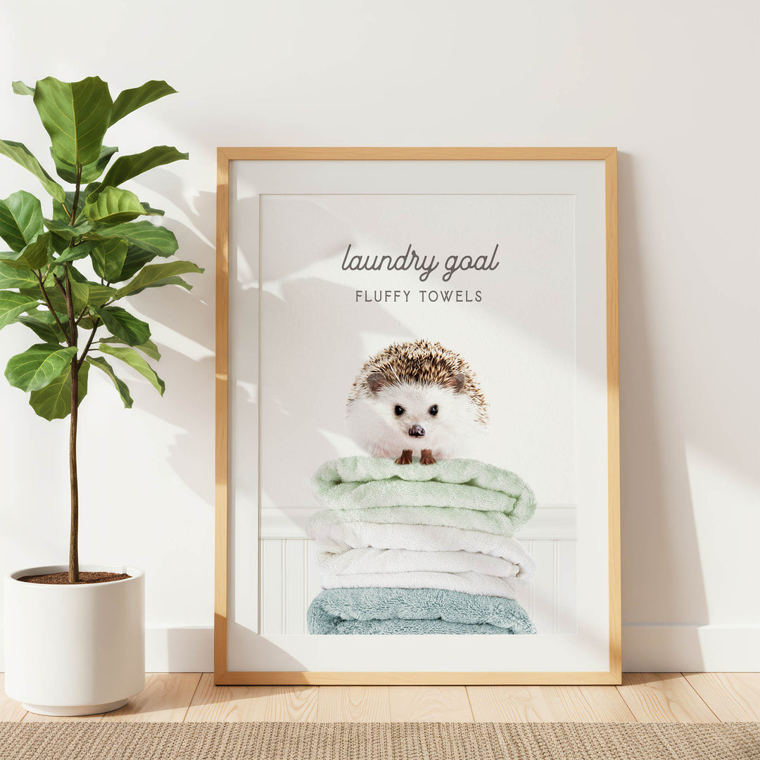 A framed poster with a hedgehog sitting atop a stack of fluffy towels, accompanied by the text "laundry goal FLUFFY TOWELS".