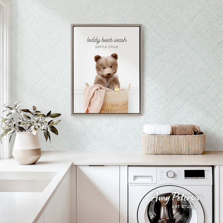 a framed photo of a teddy bear sitting in a basket, with the caption "teddy bear wash gentle cycle" above it. The photo is displayed on a wall above a white countertop, which has a potted plant and a basket of towels on it. There is also a washing machine visible in the background.