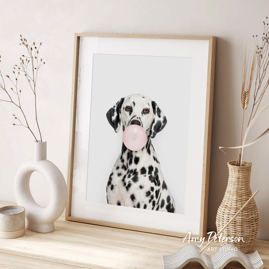 A Dalmatian dog is sitting in front of a framed picture, blowing a bubble with a pink bubble gum bubble. The dog is surrounded by decorative elements, including a white vase and a woven basket.