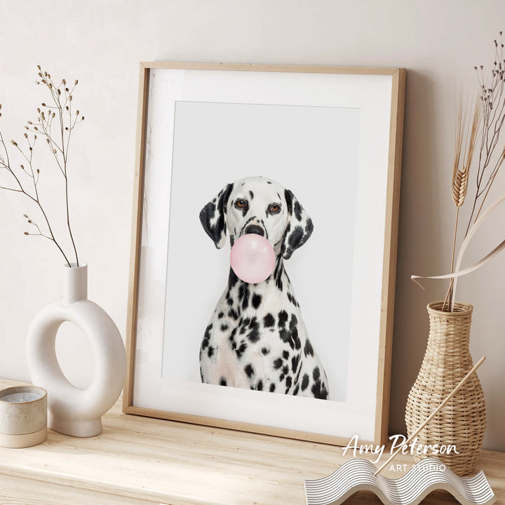 A Dalmatian dog is sitting in front of a framed picture, blowing a bubble with a pink bubble gum bubble. The dog is surrounded by decorative elements, including a white vase and a woven basket.