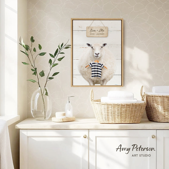 a framed picture of a sheep wearing a striped shirt, hanging on a wall above a white basket with towels. The wall has a patterned wallpaper, and there is a vase with green leaves and a bottle of soap on a white countertop.
