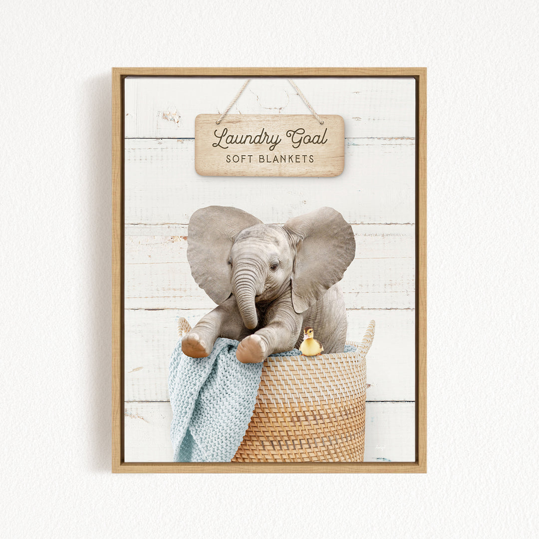 A framed image of a baby elephant sitting in a woven basket, with a sign above it that says "Laundry Goal Soft Blankets".