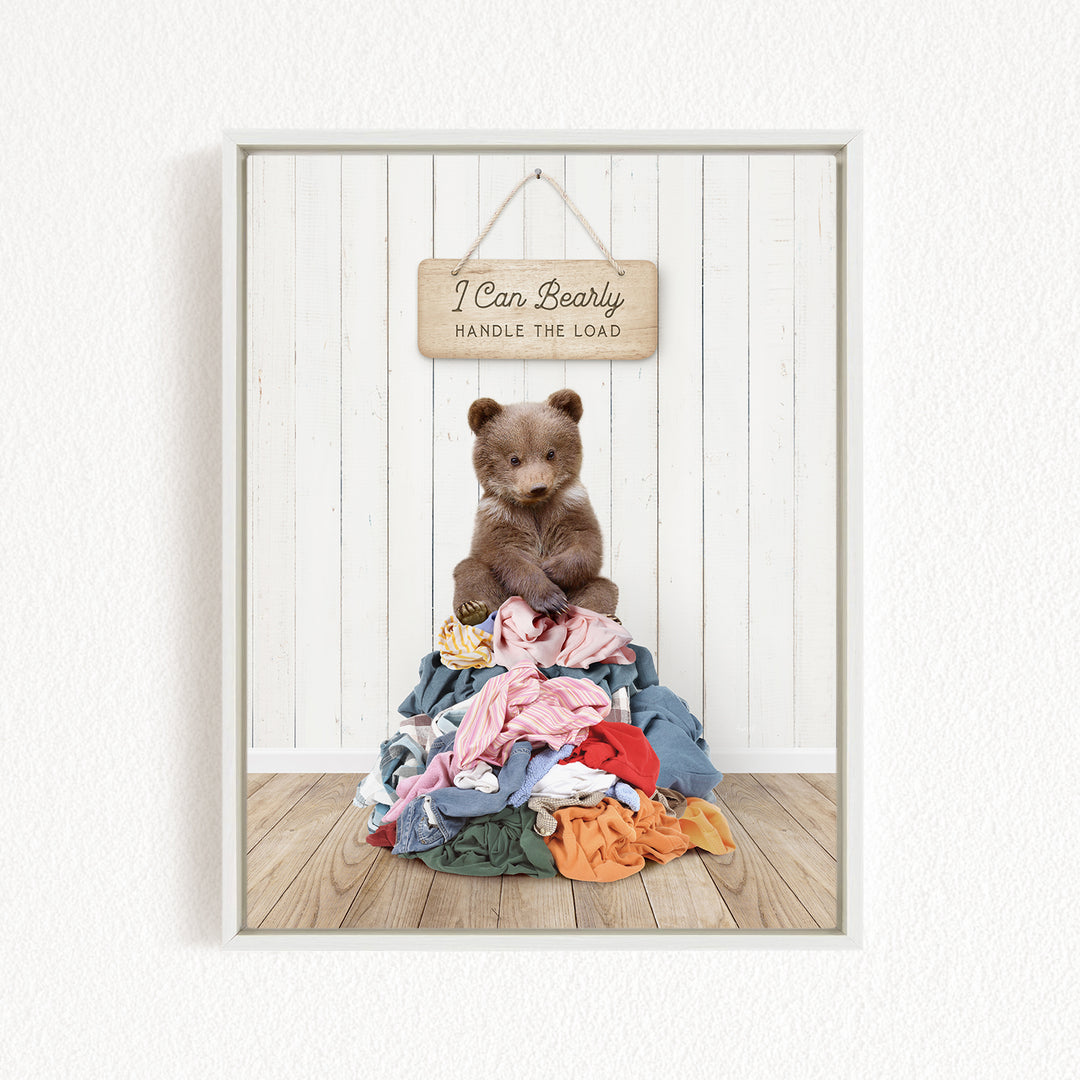 A teddy bear sitting atop a pile of clothes, with a wooden sign above that reads "I Can Bearly Handle the Load".