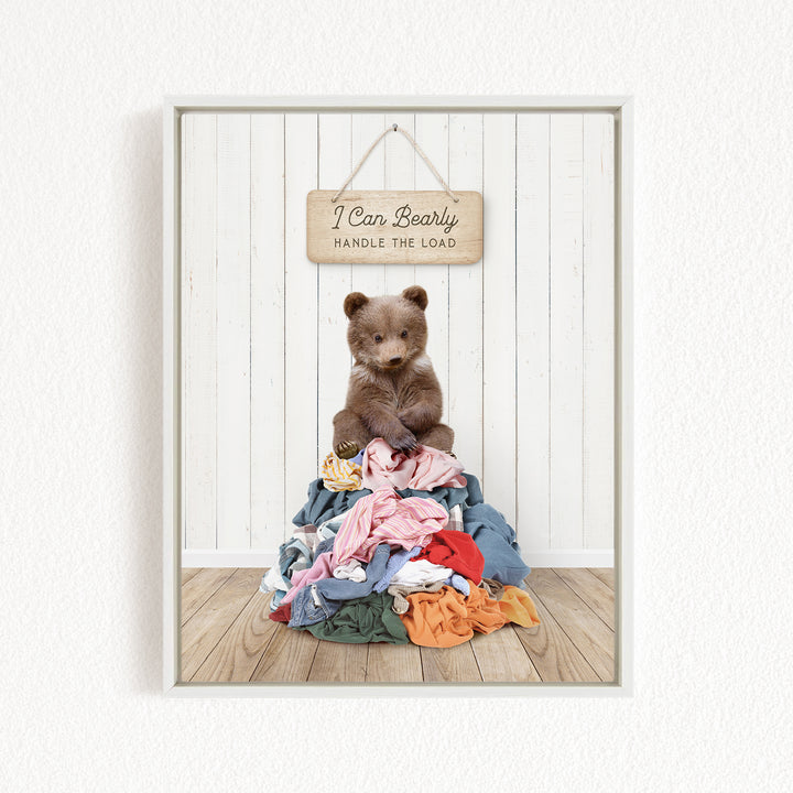 A teddy bear sitting atop a pile of clothes, with a wooden sign above that reads "I Can Bearly Handle the Load".