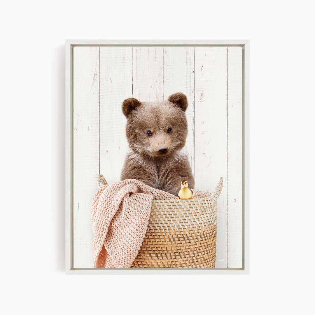 A brown bear sitting in a woven basket, with a small yellow duck nearby, against a white wooden background.