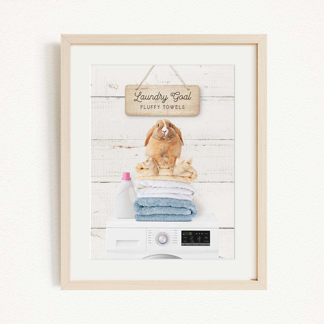 A framed image of a rabbit sitting on a stack of folded towels, with a sign above that reads "Laundry goal Fluffy towels".