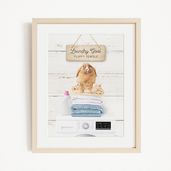 A framed image of a rabbit sitting on a stack of folded towels, with a sign above that reads "Laundry goal Fluffy towels".