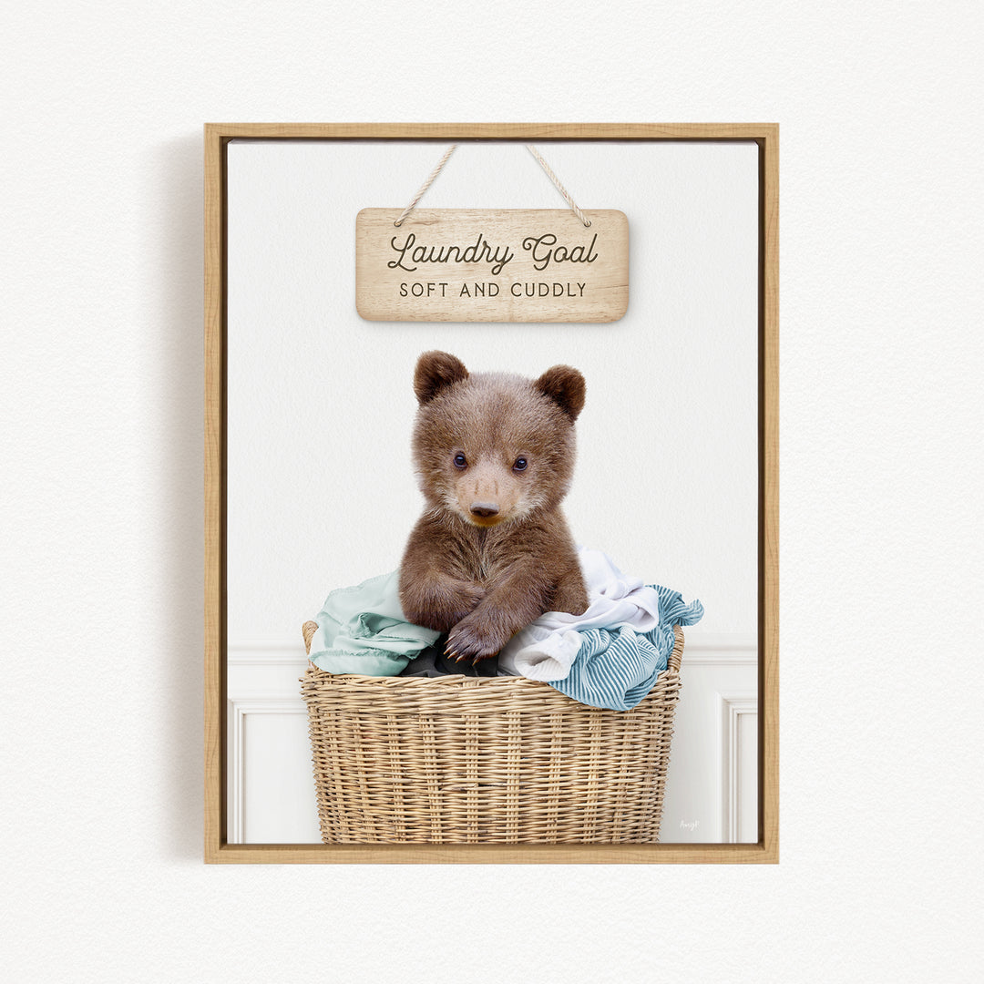 A cute brown bear sitting in a wicker laundry basket, with a wooden sign above that says "Laundry Goal Soft and Cuddly".