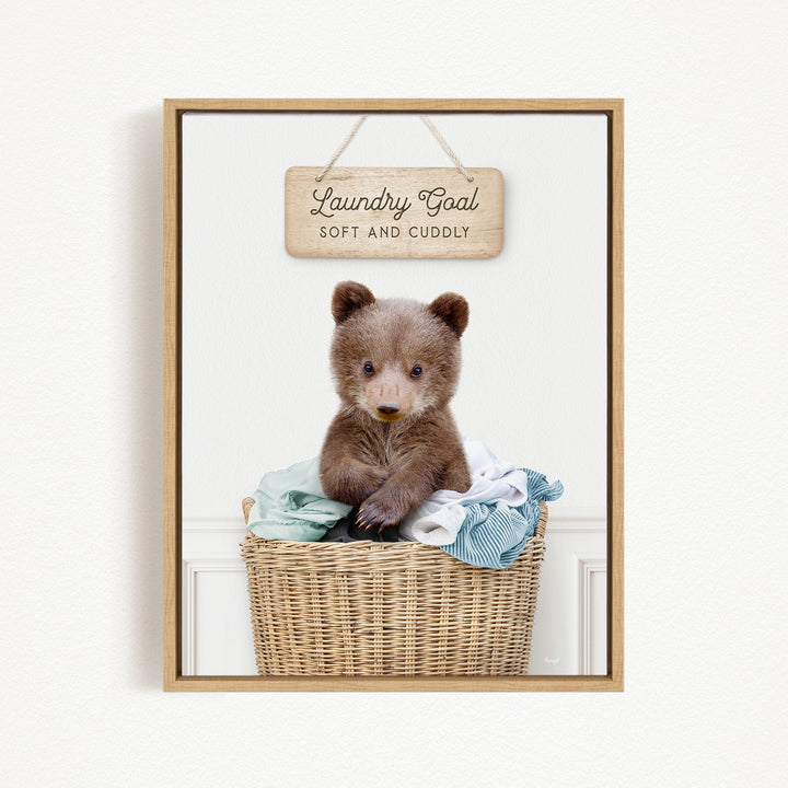 A cute brown bear sitting in a wicker laundry basket, with a wooden sign above that says "Laundry Goal Soft and Cuddly".