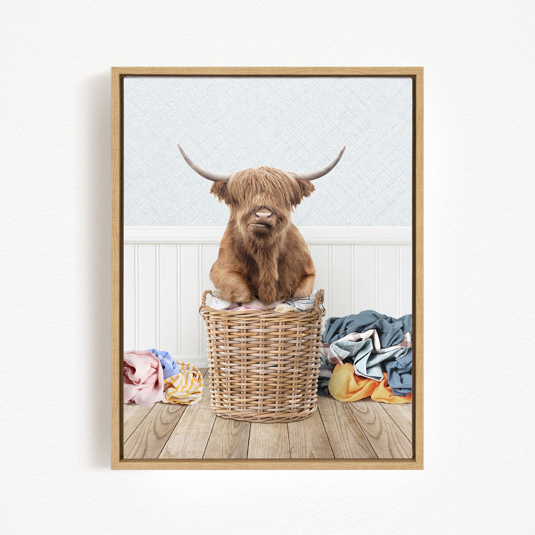 A Highland cow with long horns is sitting in a wicker basket filled with clothes, placed on a wooden floor.