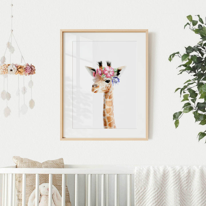 A framed picture of a giraffe with a flower crown hangs on a white wall, accompanied by a hanging mobile and a plant.