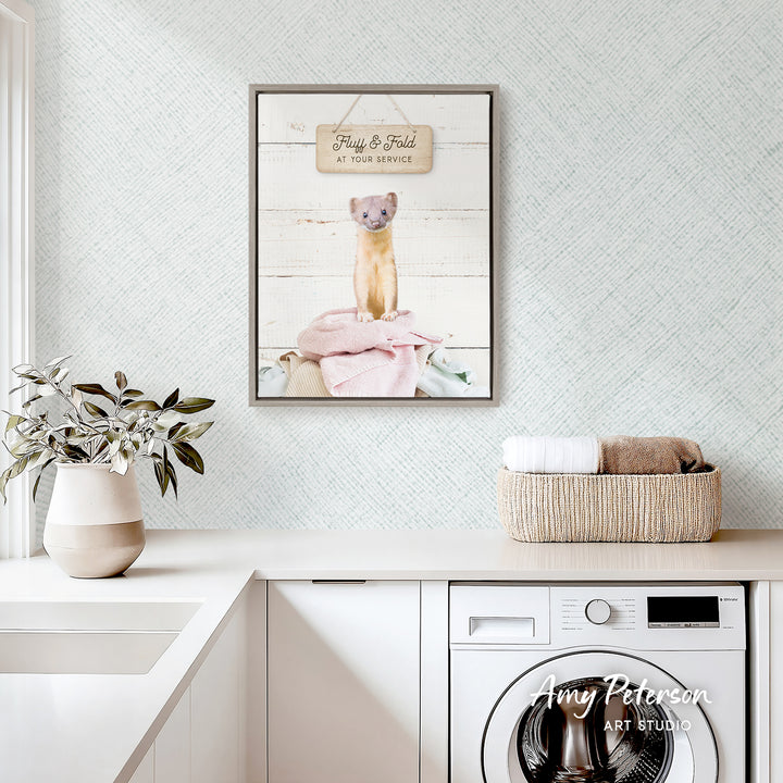 a framed picture of a cat sitting on a towel, hanging on a wall above a laundry basket and a white sink.