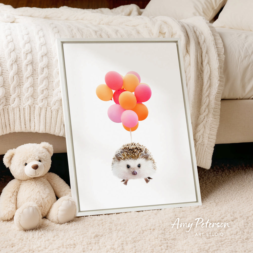 A framed print of a hedgehog holding colorful balloons is displayed on a white surface, with a teddy bear sitting next to it.
