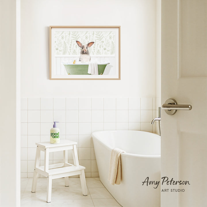 A framed picture of a rabbit sitting in a bathtub is hanging on a white tiled wall above a white bathtub in a bathroom.