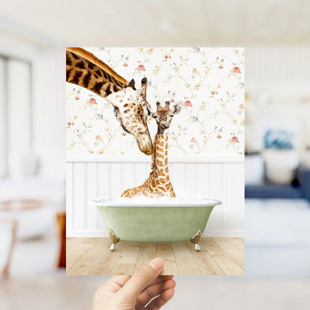A hand holds up a framed photograph of two giraffes sitting in a green bathtub, with a floral wallpaper in the background.