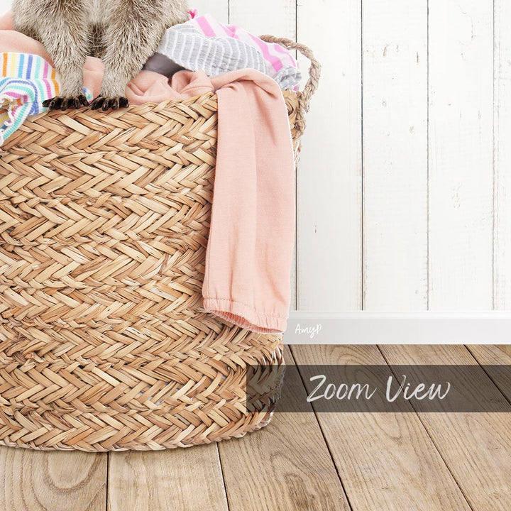 A woven laundry basket filled with clothes, including a pink garment, sits on a wooden floor against a white wall.