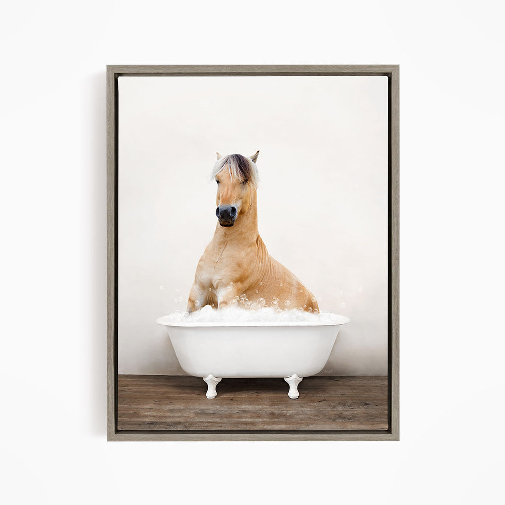 A brown horse sitting in a white bathtub filled with bubbles, with a wooden floor beneath it.