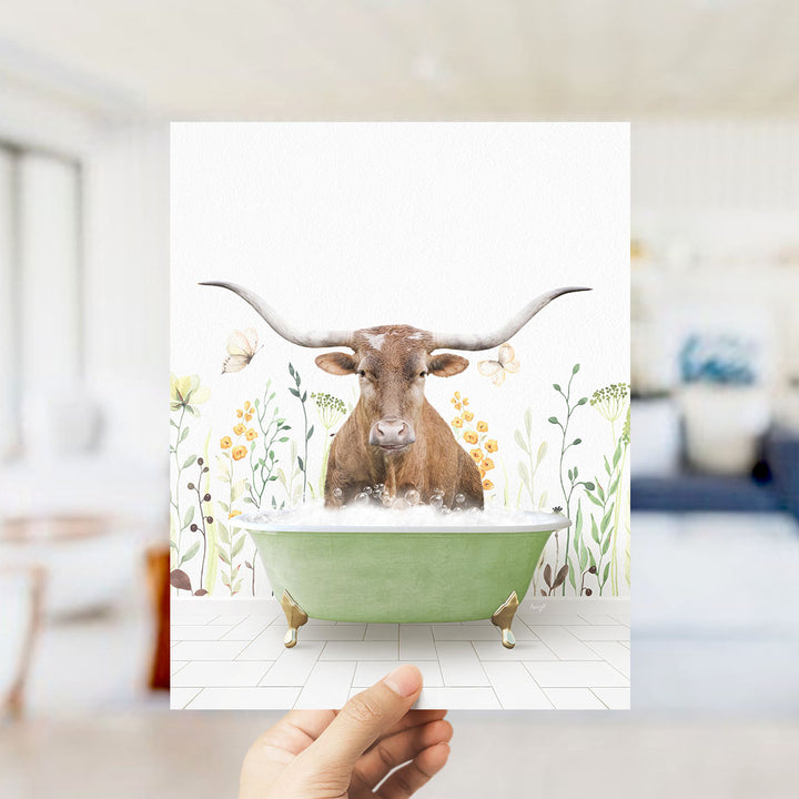 A hand holds up a framed image depicting a brown bull with large horns sitting in a green bathtub filled with water, surrounded by flowers and plants.